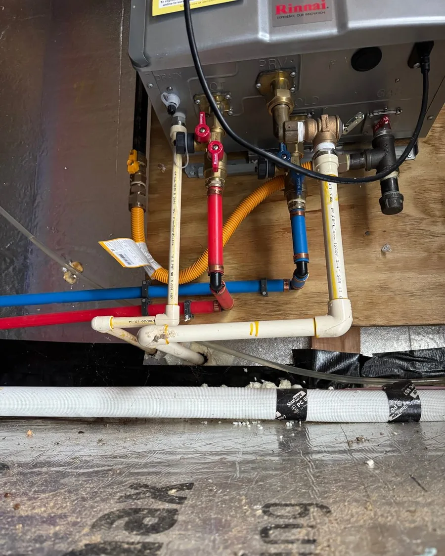 The underside of a tankless unit in a crawlspace: red and blue service valves, a pressure relief valve, white plastic drain lines and a yellow flexible gas connector