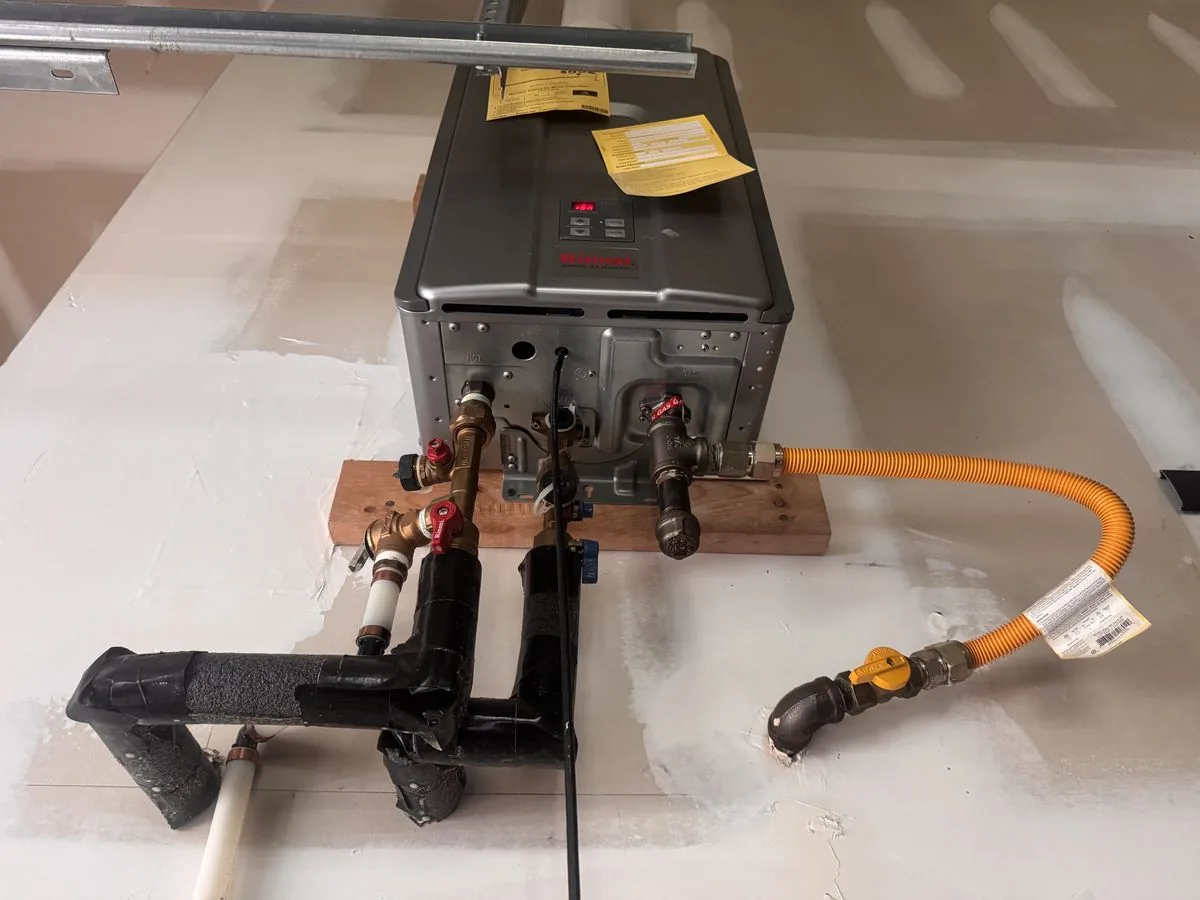 Looking up at the bottom of a tankless water heater: water service valves, black pipe insulation, and a flexible gas connector running from a yellow-handled shutoff valve