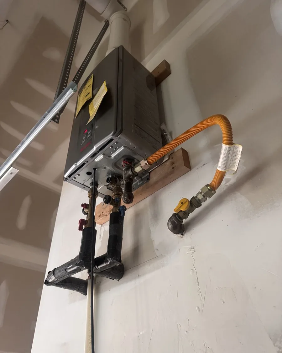 A tankless water heater mounted high on a garage wall on wood blocking, with an orange flexible gas connector and insulated water lines below it