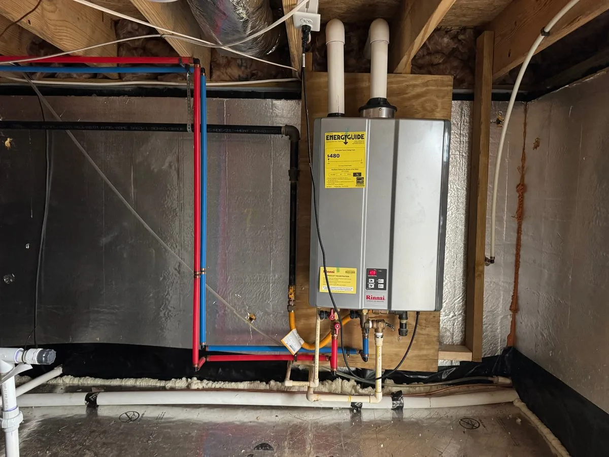 A tankless water heater hung on a plywood panel in a crawlspace lined with foil-faced foam board, with red and blue water lines running along the wall