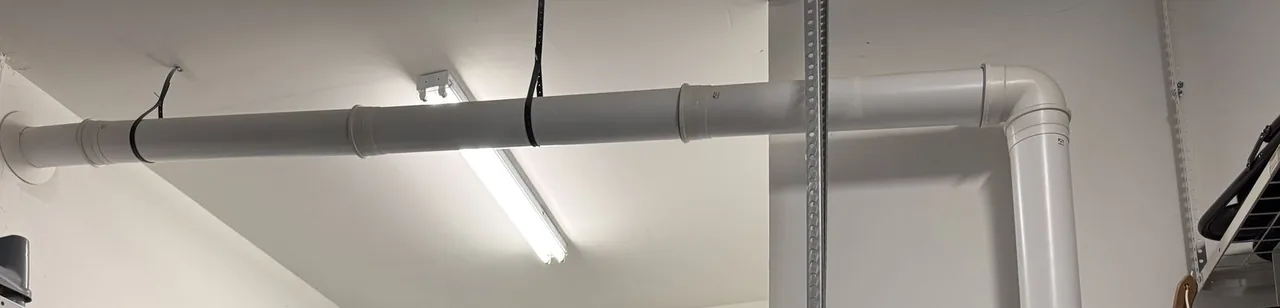 A white vent pipe hung from straps along a garage ceiling, running from a tankless water heater to an outside wall