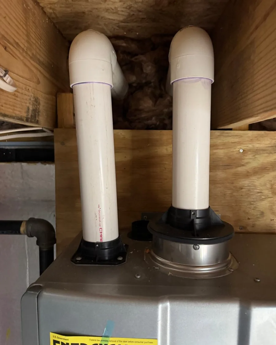 Two white plastic vent pipes rising from the top of a tankless unit and turning into the floor framing above